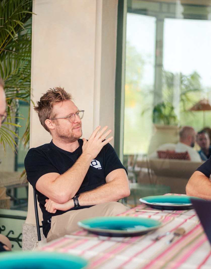 Three colleagues talking across a lunch table, palms and daylight behind them