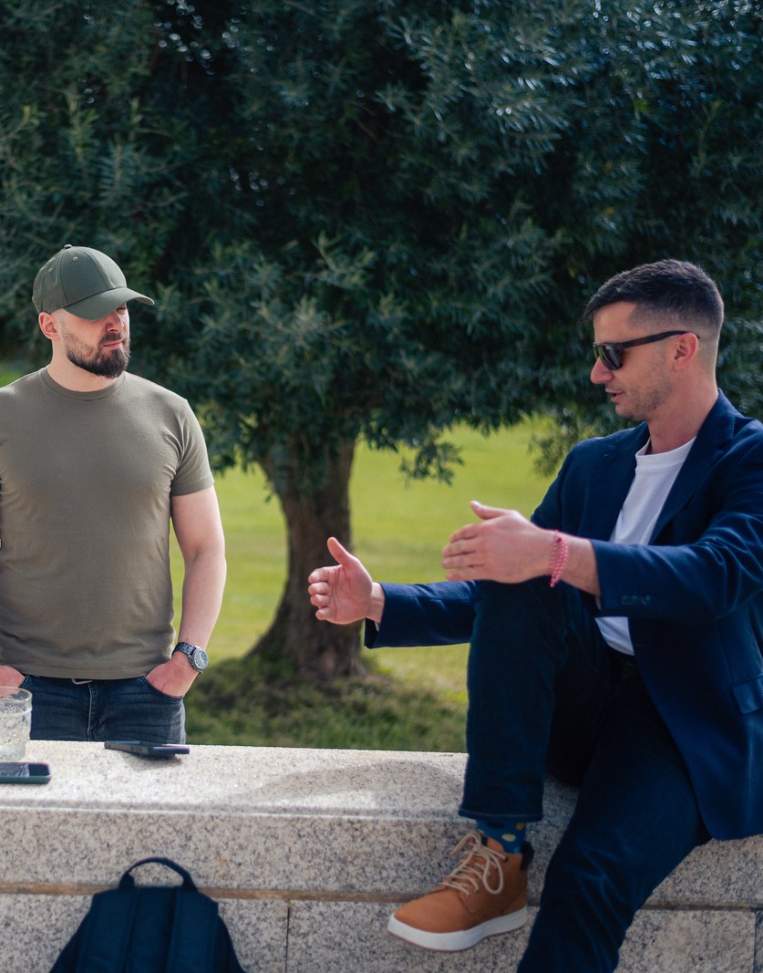 Two colleagues talking across a low stone wall under an olive tree
