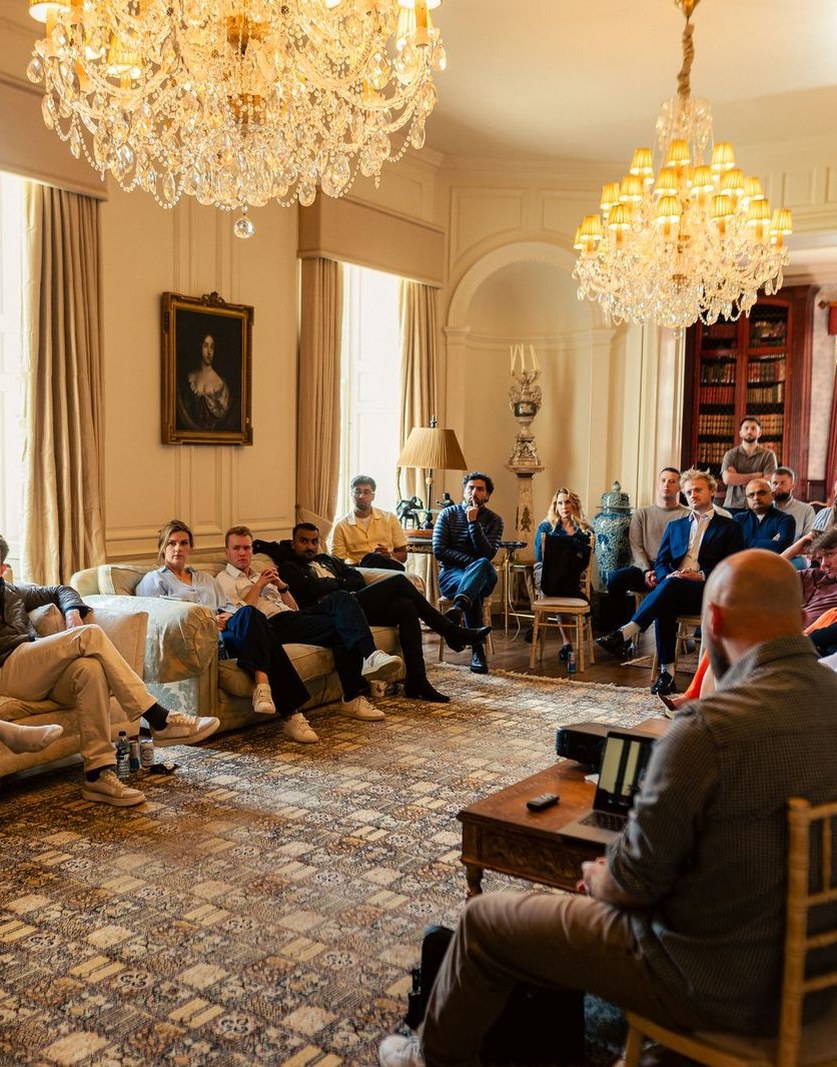 Guests seated on sofas under two chandeliers in a panelled drawing room, listening to a speaker