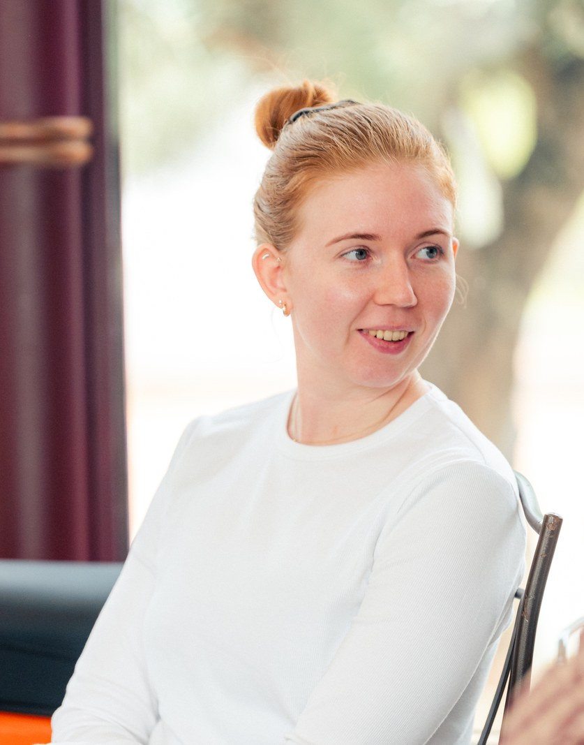 A colleague in a white top listening in a conversation, olive trees behind the glass
